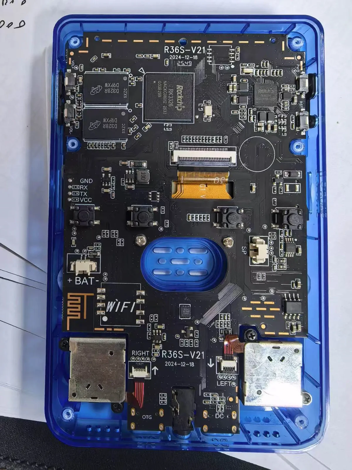 Photograph of an R36S V21 main board seen through a transparent blue shell; the silkscreen reads R36S-V21 and 2024-12-18, with WIFI pads marked on the left