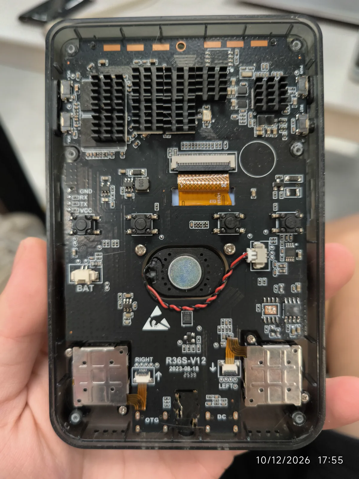 V12 board, dated 2023. The oldest of the three revisions, held in the hand. The date burned into the bottom right of the frame, 10/12/2026, is the camera's, not the board's. The silkscreen prints R36S-V12 and 2023-08-18 in the same position the V30 uses, with 2603 below it, RIGHT and LEFT beside the shoulder mounts, and OTG and DC along the bottom edge. Where the V30 photograph shows one gold heat spreader, this board carries two black finned ones. Same layout family, two years apart, in a black shell — which is the point: the shell is not the revision.