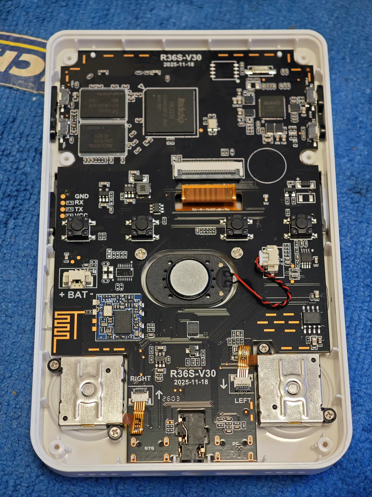 V30 board, shell off. A white unit with the back shell off. The silkscreen reads R36S-V30 and 2025-11-18 twice: along the top edge and again between the shoulder mounts, with RIGHT and LEFT printed beside those mounts and OTG and DC along the bottom edge. GND, RX, TX and VCC are labelled at mid-left, next to a + BAT marking at the battery connector. Two memory packages sit at the top left beside the Rockchip module, the round speaker and its twisted two-wire lead sit in the middle of the board, and a gold finned heat spreader covers the right-hand side.