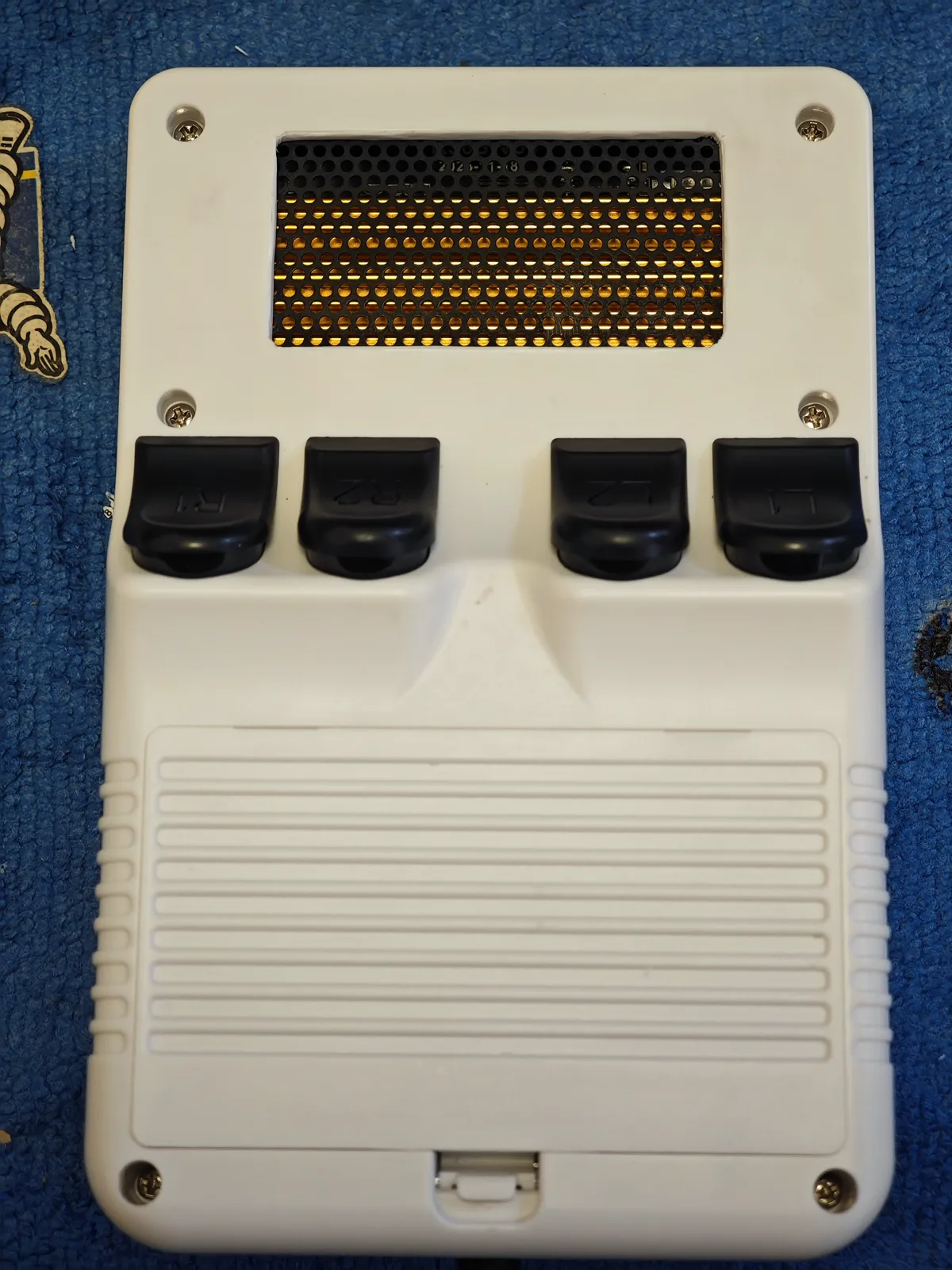 The back cover, opened out. The white back cover from the outside — the same cover as the frame below it. A rectangular opening was cut in the shell above the shoulder buttons, and a black perforated sheet sits in that opening; a gold honeycomb surface shows through it from the board behind. Below the opening are the four shoulder buttons, each carrying a moulded marking, and the ribbed grip runs down the rest of the cover. This shell is screwed together. The opening is a modification we made; it is not how the cover arrives.