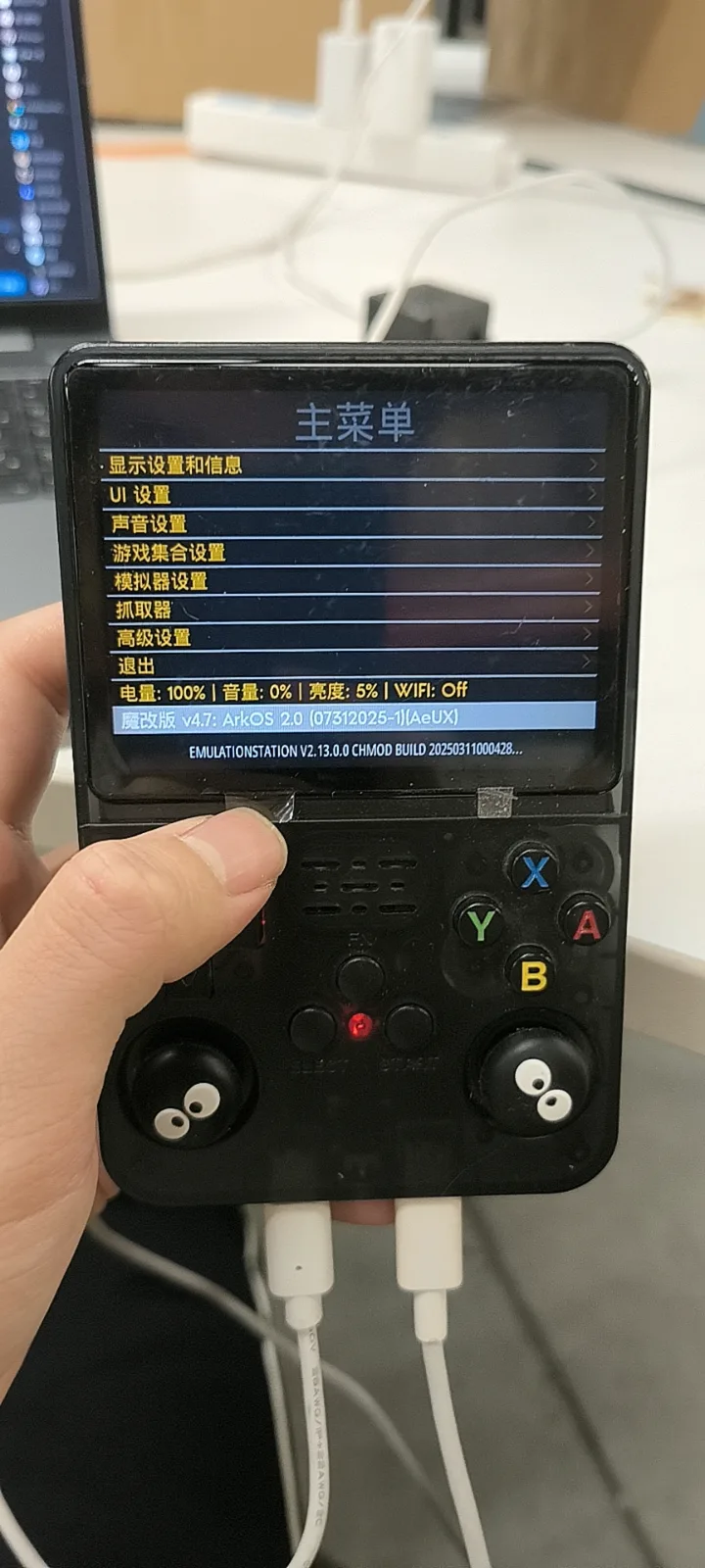 Photograph of an R36S held in one hand, showing a Chinese-language main menu; the highlighted line reads 魔改版 v4.7: ArkOS 2.0 (07312025-1)[AeUx] and the line below it reads EMULATIONSTATION V2.13.0.0 CHMOD BUILD 20250311000428